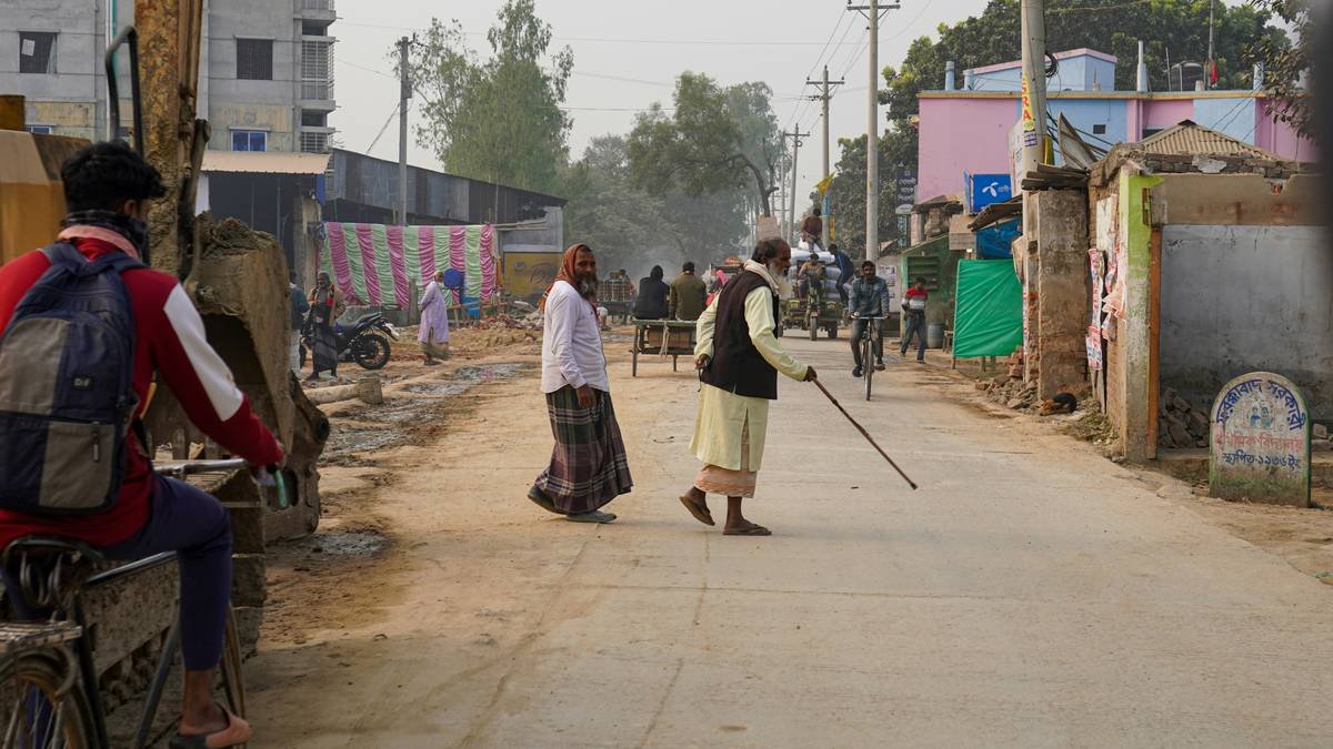 A busy street scene in a rural town showing the intersection of daily life and infrastructure development. An elderly man walks with a cane and pedestrians navigate the road alongside an excavator, while shops and local businesses bustle in the background.