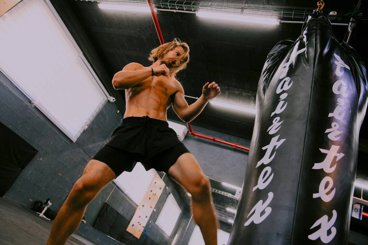 The image shows a shirtless individual in black shorts engaged in a boxing workout at a gym. The person is in a fighting stance, preparing to punch a large black punching bag with the word "Fairtex" written on it. The gym setting includes industrial lighting and a climbing wall in the background, indicating a versatile training environment. This image is relevant for illustrating intense physical training and fitness routines, highlighting the importance of strength and conditioning in sports.